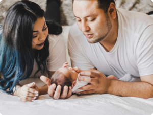 Two adults look down lovingly at a newborn baby lying between them on a white, crinkled surface.