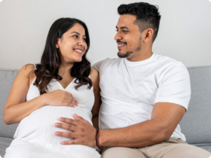 A pregnant person in a white dress and a person in a white t-shirt sitting close together on a gray couch, looking at each other and smiling.
