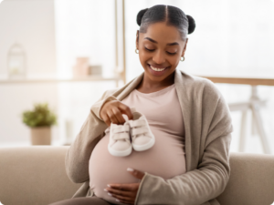 A pregnant person holds a pair of baby booties over their belly while smiling.