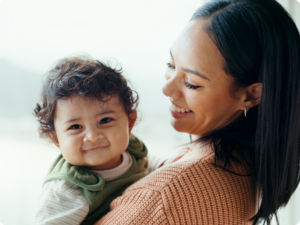 A smiling baby wearing a green hooded vest is held by a smiling person wearing a peach-colored sweater.