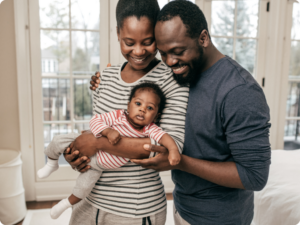 A smiling couple holds a baby between them. The baby is wearing a striped onesie.