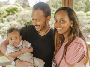 A man holds a baby while a woman smiles next to them in an outdoor setting.