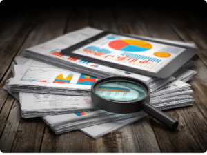 A tablet displaying charts and graphs rests on a stack of papers, with a magnifying glass placed over some of the documents on a wooden desk.
