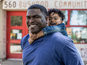 A smiling person gives a child a piggyback ride in front of a building with "560 BUSHROD COMMUNITY" visible above red doors.