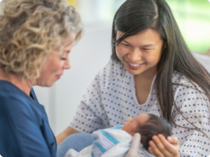 A nurse in blue scrubs and a patient in a hospital gown smile as they look down at a newborn baby wrapped in a striped blanket.
