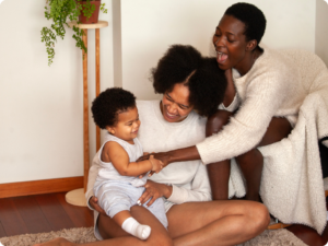 Two adults and a baby are sitting on the floor interacting and laughing.
