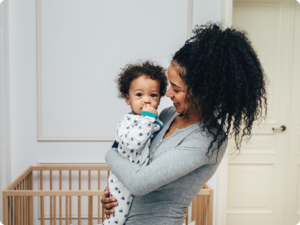 A person holds a baby in front of a crib. The baby is wearing pajamas with a star pattern and has their fingers in their mouth.