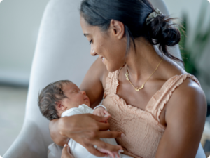 A person cradles a newborn baby, looking down at them with a gentle smile. The baby is swaddled in white, and the person wears a peach-colored top and a delicate necklace.