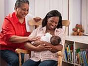 A woman smiling and holding an infant with another women sitting behind them smiling. 