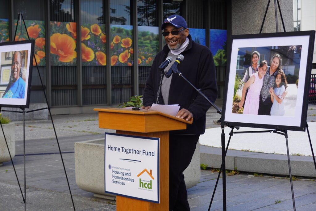 Image of a gentleman speaking at a podium at an outdoor press conference. He wears a blue cap, sunglasses, and a black jacket. 