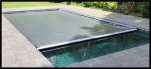 Automatic pool cover partially closed over a rectangular in-ground swimming pool with surrounding stone deck